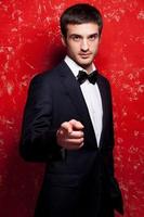 Confident and stylish. Handsome young man in suit and bow tie pointing you while standing against red background photo
