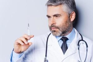 Doctor with syringe. Confident mature doctor holding syringe and looking at it while standing against grey background photo