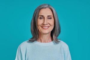 Headshot of mature beautiful woman looking at camera and smiling while standing against blue background photo