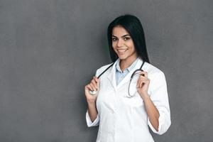 You can absolutely trust me Attractive young female doctor in white lab coat keeping arms crossed and smiling while standing against grey background photo