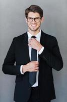 Everything should be perfect. Happy young businessman adjusting his necktie and looking at camera while standing against grey background photo