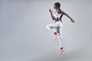 toda la longitud de una joven africana segura de sí misma vestida con ropa deportiva saltando contra un fondo gris foto