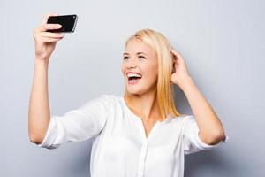 Expert in photography. Cheerful young blond hair woman holding mobile phone and making photo of herself while standing against grey background