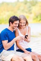 Posting their pictures online. Beautiful young loving couple sitting at the quayside and looking at mobile phone together photo