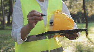 trabajador asiático inteligente o ingeniero civil masculino con casco de seguridad protector y chaleco reflectante que usa tableta digital para planificar proyectos y verificar el dibujo arquitectónico en el sitio de construcción. video