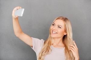 Selfie time Cheerful young woman making selfie on her smart phone while standing against grey background photo