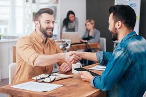 Well done Two confident young men shaking hands and smiling while sitting at the desk in office with two people working in the background photo