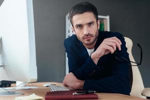 Need an expert advice Confident young man carrying eyeglasses and looking at camera while sitting at his working place in office photo