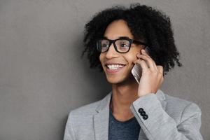 Having some great news Cheerful young African man talking on mobile phone and smiling while standing against grey background photo