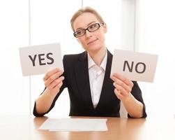 Maybe yes Thoughtful mature woman in formalwear sitting at the table and holding papers with yes and no on them photo