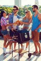 Cheers to friends Full length of fourjoyful young people clinking glasses with beer and barbecuing while standing on the roof photo