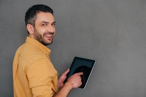 How may I help you  Rear view of handsome mature man holding digital tablet and looking over shoulder with smile while standing against grey background photo