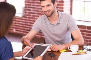 Let me show you all the features. Two cheerful people in casual wear sitting face to face at the table while man pointing digital tablet and smiling photo