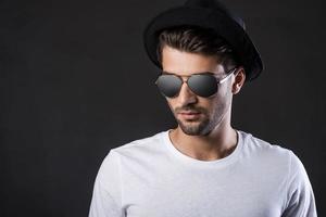 confiado y guapo. apuesto joven con gafas de sol y fedora de pie contra el fondo negro foto