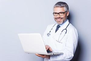 Cheerful doctor with laptop. Confident mature doctor working on laptop while standing against grey background photo