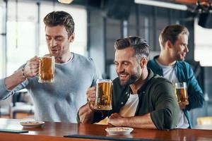 Group of cheerful young men in casual clothing enjoying beer and communicating photo