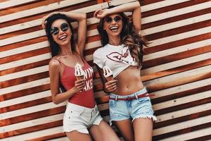 nada mejor que un helado. dos atractivas mujeres jóvenes sonriendo y comiendo helado mientras están de pie contra la pared de madera al aire libre foto