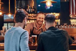 camarero alegre que sirve cerveza a los jóvenes mientras está de pie en el mostrador del bar en el pub foto