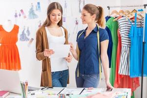 Discussing new fashion look. Two attractive female designers discussing something while looking at the paper photo