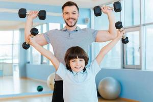 As strong as my father. Happy father and son exercising with dumbbells and looking at camera while both standing in health club photo