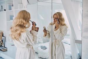 Attractive young woman in bathrobe doing make-up while looking at her reflection in the mirror photo