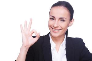 gesticulando bien signo. mujer joven feliz en ropa formal gesticulando signo ok y sonriendo mientras está de pie aislado en blanco foto