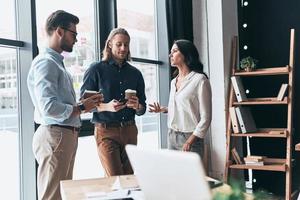 Coffee break. Young colleagues in smart casual wear discussing something and holding disposable cups while standing in the office photo
