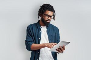 Thoughtful young African man in eyewear using digital tablet while standing against grey background photo