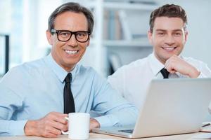 Two professionals at work.Two cheerful business people in formalwear working on laptop and smiling at camera while sitting at working place photo