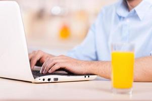 Man working on laptop. Cropped image of man in shirt working on laptop while sitting in the kitchen with glass of orange juice on foreground photo