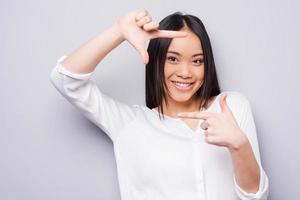 Focus on me. Beautiful young Asian woman looking at camera and gesturing finger frame while standing against grey background photo