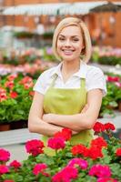 I have the best job in the world Attractive young blond hair woman in apron keeping arms crossed and smiling while standing in a greenhouse with flowers all around her photo