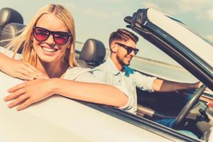 Feeling relaxed and free. Happy young woman leaning at the vehicle door and looking at camera while her boyfriend sitting near and driving convertible photo