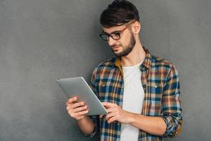 Enjoying his digital tablet. Confident young man holding his digital tablet and looking at it while standing against grey background photo