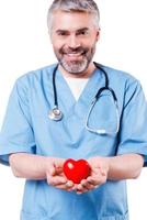 Taking good care of your heart. Confident mature cardiology surgeon holding heart shape toy and smiling while standing isolated on white photo