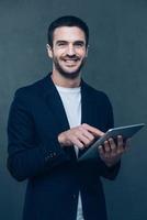 Enjoying his new digital tablet. Cheerful young man holding digital tablet and smiling while standing against grey background photo