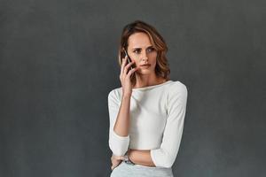 Good talk with client. Beautiful young woman in smart casual wear talking on her smart phone while standing against grey background photo