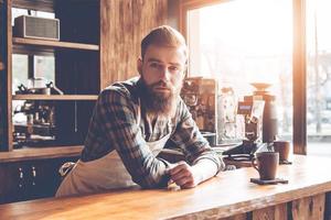 ¿Cómo puedo ayudarte, joven barbudo con delantal mirando a la cámara mientras se inclina hacia el mostrador del bar en el café? foto