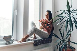 Attractive young woman in cozy pajamas using smart phone and enjoying her bouquet of tulips while resting on the window sill at home photo