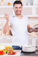 sabía tan bien un joven apuesto probando la sopa de la sartén y sonriendo mientras estaba de pie en la cocina foto