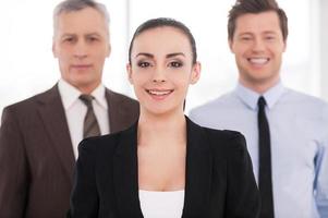 Confident in her team. Attractive young woman in formalwear looking at camera and smiling while his colleagues standing behind her photo