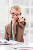 I choose you Cheerful little girl in glasses and formalwear sitting at the table and pointing you photo