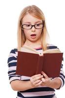 No way Surprised little girl reading book and keeping mouth open while standing isolated on white photo