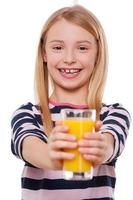 Take a juice Cheerful little girl stretching out hands with glass of orange juice and smiling at camera while standing isolated on white photo