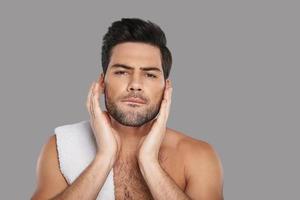 His skin needs good care. Handsome young man looking at camera and touching his face while standing against grey background photo