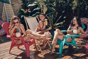 Attractive young women in swimwear drinking cocktails and smiling while relaxing outdoors photo