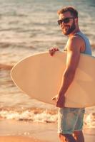 Loving summer. Side view of young man holding skimboard and smiling at camera while walking along the beach photo