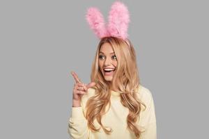 Surprised young smiling woman in pink bunny ears pointing copy space while standing against grey background photo