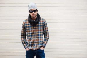 Confident in his style. Handsome young man in hat and sunglasses holding hands in pockets and looking at camera while standing against brick wall photo