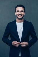 Buttoning his jacket. Cheerful young man adjusting his jacket and smiling while standing against grey background photo